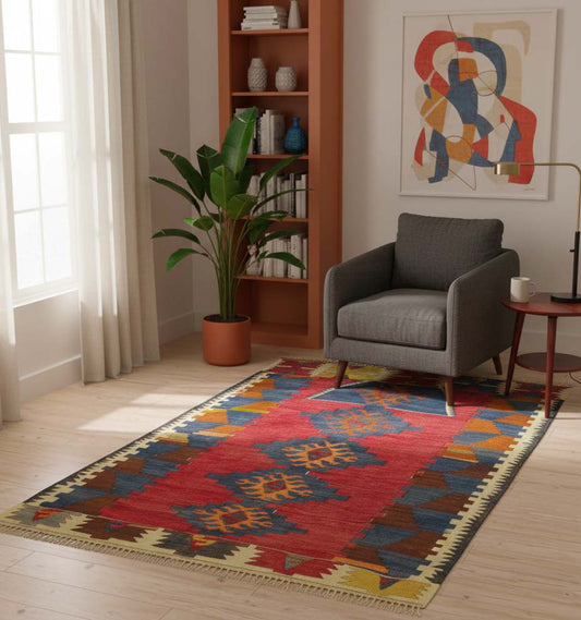 Tapis de salon en laine rouge avec larges bordures et motifs bleus (150 x 102 cm) mis en situation devant un fauteuil gris et une étagère terre cuite