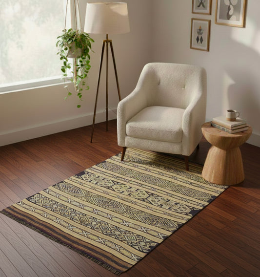 Petit tapis de salon en laine aux motifs géométriques ocre et noir (115 x 92 cm) mis en situation devant un fauteuil bouclé blanc avec une table d'appoint en bois