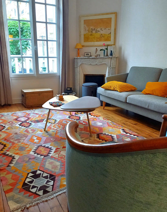 Kilim ancien d'Anatolie aux grands motifs géométriques orange, ocre et brun posé dans un salon vintage avec cheminée en marbre et mobilier scandinave à Paris.