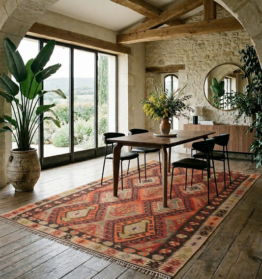 Grand tapis Kilim en laine aux motifs géométriques oranges sous une table de salle à manger en bois dans un décor rustique chic avec murs en pierre.