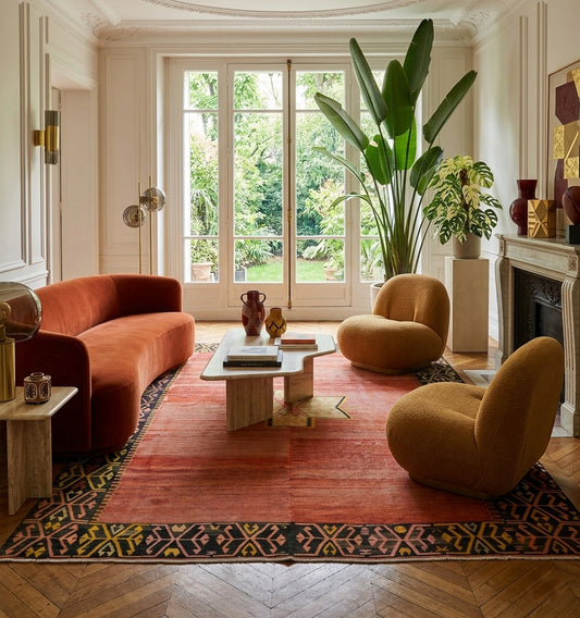 Grand tapis kilim ancien d'Anatolie aux teintes terre de Sienne, contrastant avec un canapé galbé en velours orange et des fauteuils bouclés dans un salon luxueux à Paris.