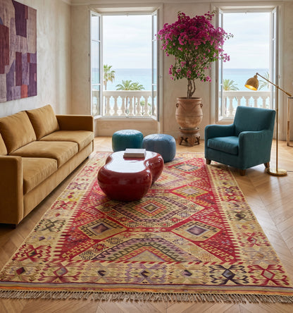 Grand tapis kilim ancien d'Anatolie aux motifs géométriques complexes et teintes chaudes, structurant un vaste salon chic face à la mer à Nice. Ce tissage artisanal d'exception crée un contraste saisissant avec un canapé ocre, un fauteuil bleu canard et une table design laquée rouge.