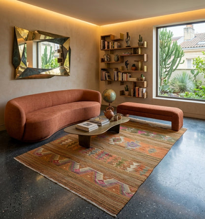 Kilim ancien aux tons pastel et terre cuite, harmonisant un salon au design organique avec canapé courbe, miroir sculptural doré et sol en béton ciré pour une ambiance chic et apaisante.