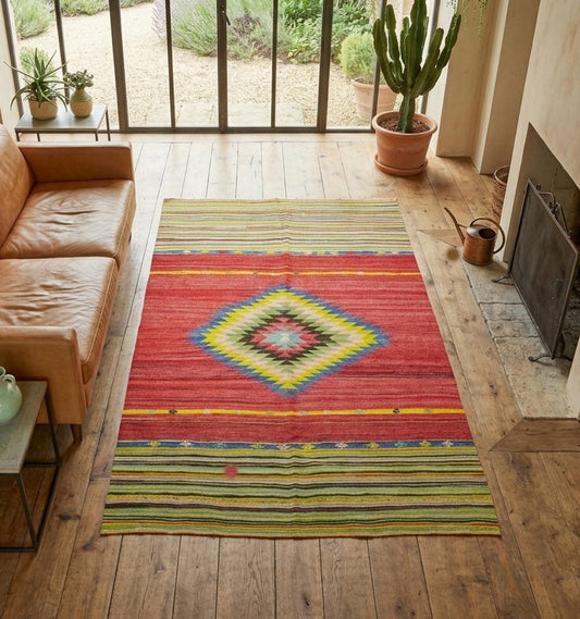 Kilim anatolien aux rayures multicolores et motif central en diamant, présenté dans un salon élégant avec une vue sur un jardin avec des cailloux, et un canapé en cuir marron clair. 