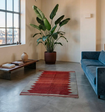 Tapis Kilim Balikesir vintage rouge et gris en laine dans un salon moderne avec sol en béton ciré, canapé bleu et plante verte