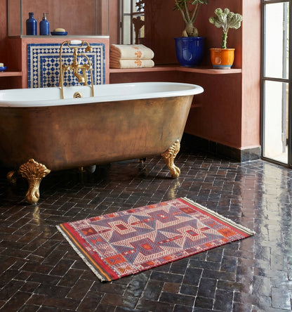 Kilim ancien de Kozak, Anatolie, motifs géométriques bleu, rouge et crème tissé main en laine naturelle, devant une baignoire dorée dans un riad à Marrakech.