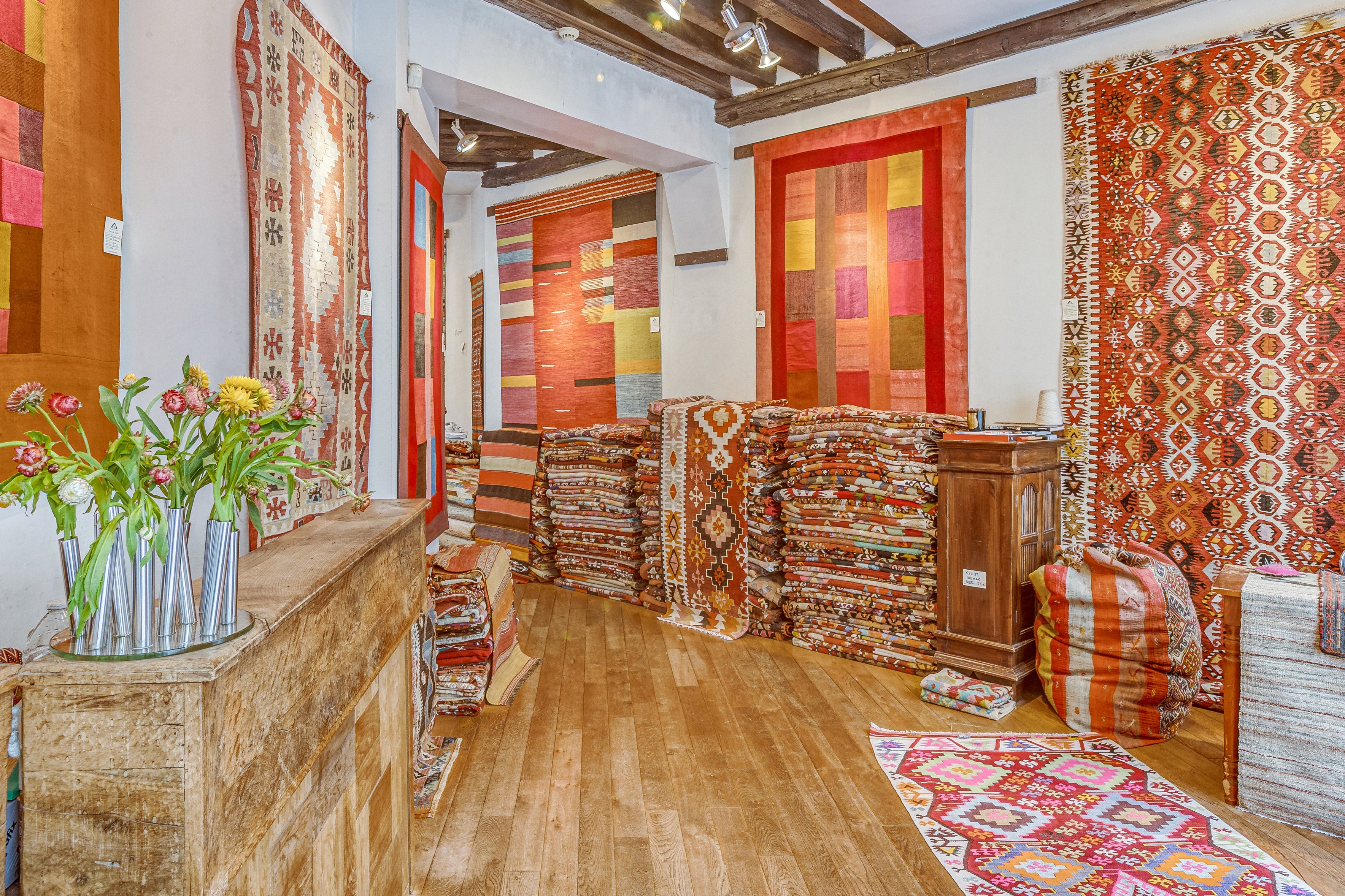 Vue d'ensemble chaleureuse et lumineuse de l'intérieur de notre magasin de tapis à Paris, mettant en valeur une large collection de kilims d'Anatolie et de tapis persans tissés à la main. Au premier plan, un comptoir en bois patiné avec un vase de fleurs printanières (œillets) et un terminal de paiement. Les murs sont décorés de grands tapis aux motifs géométriques vibrants (tons de rouge, d'orange, de jaune et de bleu). De hautes piles de kilims pliés sont soigneusement alignées sur un parquet en bois.