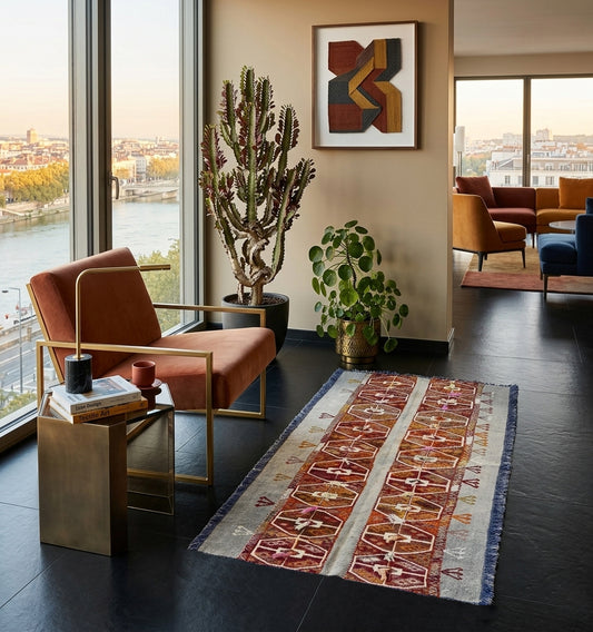 Petit tapis kilim d'Anatolie aux motifs tribaux et bordures bleues, disposé sur un sol noir moderne avec fauteuil en velours orange et vue plongeante sur la Loire à Lyon.