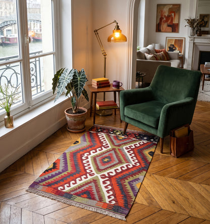 Petit tapis kilim ancien aux motifs géométriques colorés, disposé au pied d'un fauteuil en velours vert dans un appartement haussmannien près du Canal Saint-Martin à Paris. Une touche d'artisanat d'Anatolie qui réveille un coin lecture chic et vintage.