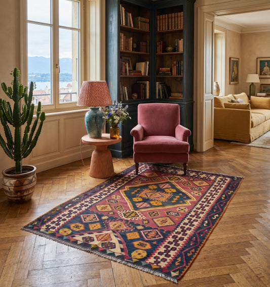 Tapis de salon en laine rouge avec un grand losange central et motifs géométriques (168 x 126 cm) mis en situation devant un fauteuil bleu avec des coussins au sol