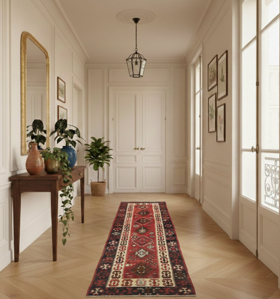 Long tapis de couloir en laine Komurgen rouge aux motifs traditionnels (343 x 72 cm) mis en situation dans une galerie haussmannienne avec parquet en point de Hongrie, murs à moulures blanches, console classique et miroir doré