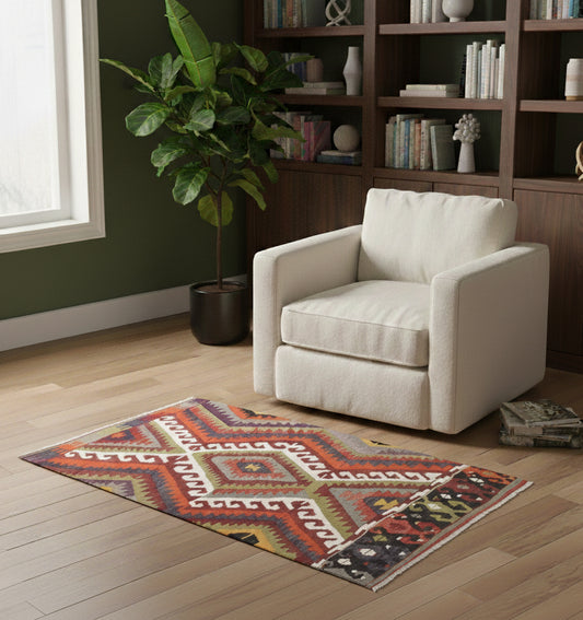 Petit tapis en laine Antalya aux motifs géométriques colorés (116 x 76 cm) mis en situation dans un coin lecture élégant avec un large fauteuil blanc, une bibliothèque en bois sombre et un mur d'accent vert forêt