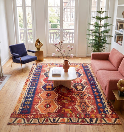 Tapis kilim ancien d'Adana, tissé main en laine par des nomades d'Anatolie, médaillons hexagonaux rouge et orange sur bordure bleu nuit, posé dans un salon à Deauville face à une maison à colombages et un balcon en fer forgé