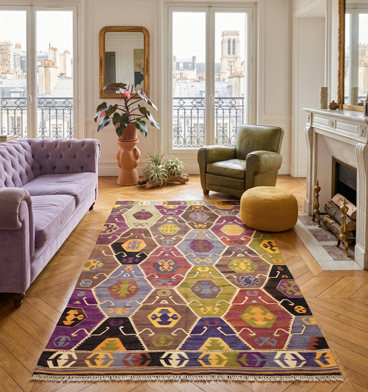Tapis kilim ancien d'Anatolie aux motifs hexagonaux colorés, structurant un grand salon haussmannien avec vue sur les toits à Paris. L'élégance du tissage artisanal traditionnel mariée à un canapé en velours parme et un mobilier chic contemporain.