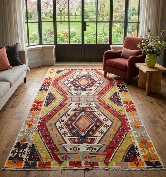 Tapis de salon en laine Avsar aux motifs géométriques traditionnels rouge, jaune et bleu (255 x 146 cm) mis en situation dans un salon rustique avec parquet en bois massif, cheminée en pierre et canapé crème.