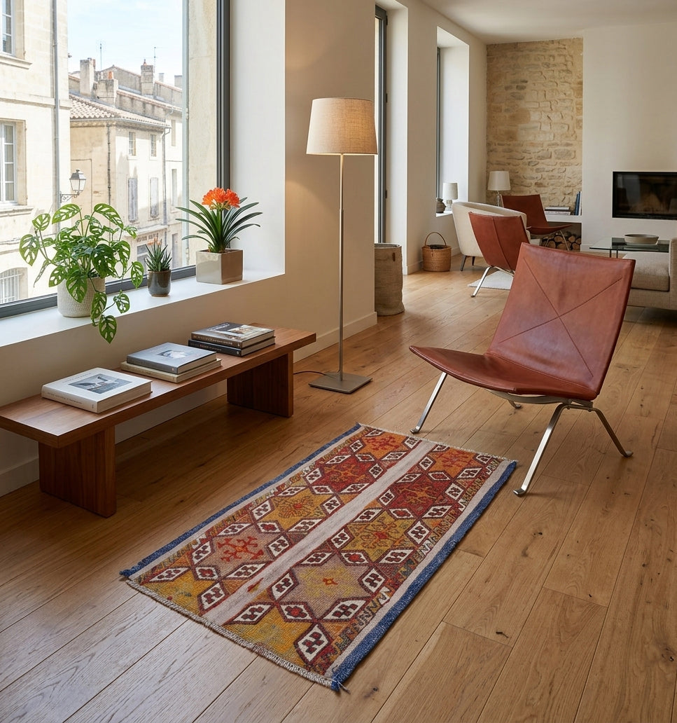 Petit tapis kilim Cuval ancien (60 ans) aux motifs géométriques tribaux, tissé main en Anatolie. Cette pièce de collection artisanale, aux tons chauds et bordures bleues, crée un contraste élégant et authentique dans un salon contemporain avec mobilier en cuir et parquet en chêne.