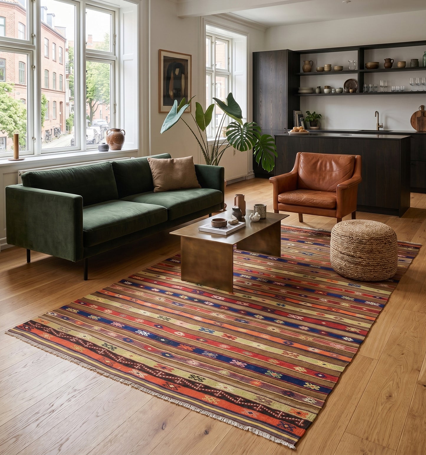 Tapis kilim Fethiye de 55 ans aux rayures multicolores et motifs tribaux, tissé à la main en Anatolie. Cette grande pièce artisanale apporte une chaleur authentique à un salon-cuisine ouvert à Nantes, créant un contraste chic avec un mobilier moderne en velours vert, un fauteuil en cuir et une décoration minimaliste.