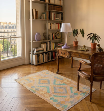 Tapis kilim tissé main aux losanges turquoise et sable, tapis contemporain d’inspiration Anatolie posé sur parquet en chevrons dans un bureau parisien face à un secrétaire en bois et une bibliothèque