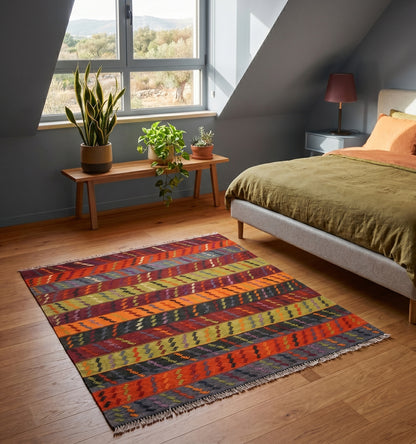 Tapis kilim Manisa ancien aux rayures diagonales multicolores, tissé à la main en laine de haute qualité. Cette pièce artisanale turque apporte une touche de couleur vive et d'authenticité à une chambre moderne mansardée, créant un contraste harmonieux avec des murs gris bleu, un parquet en bois naturel et une décoration minimaliste agrémentée de plantes vertes.
