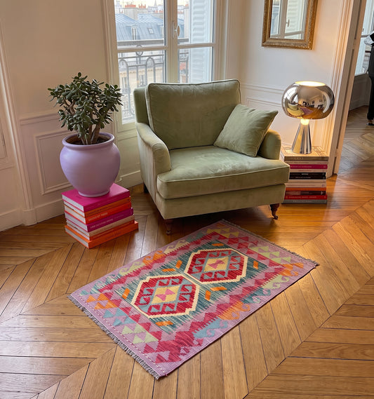 Tapis kilim tissé main aux médaillons géométriques rose et sarcelle sur fond lilas, tapis contemporain d'inspiration Anatolie posé dans un appartement haussmannien face à un fauteuil en velours sauge
