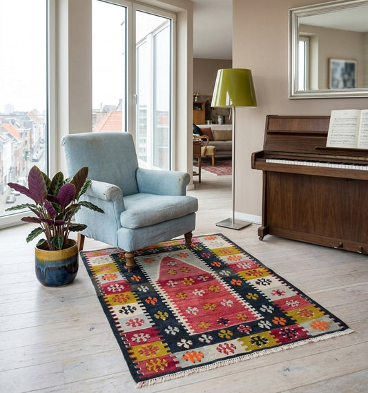 Tapis kilim tissé main au mihrab rose framboise façon tapis de prière, bordures fleuries jaune safran et bleu nuit, posé dans un salon à Anvers face à un piano droit et la vue sur les toits