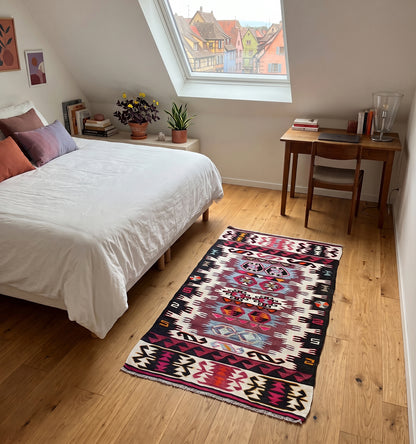 Tapis kilim tissé main aux motifs tribaux fuchsia et noir, tapis ancien posé sous velux dans une chambre à Colmar face aux maisons à colombages colorées