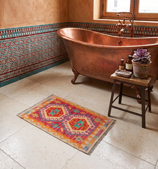 Tapis kilim tissé main aux octogones orange et fuchsia bordés de turquoise, tapis contemporain d'inspiration Anatolie posé dans une salle de bain à Marrakech face à une baignoire en cuivre et un zellige ancien