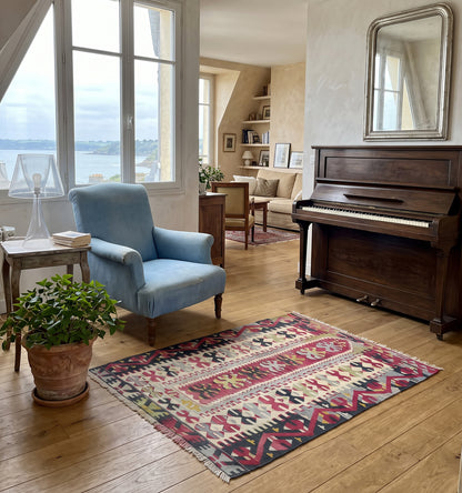 Tapis kilim tissé main aux rayures et croix grenat et vert amande, motifs géométriques hérités des tisserands d'Anatolie, posé dans un salon à Saint Malo face à un piano ancien et la vue sur la baie