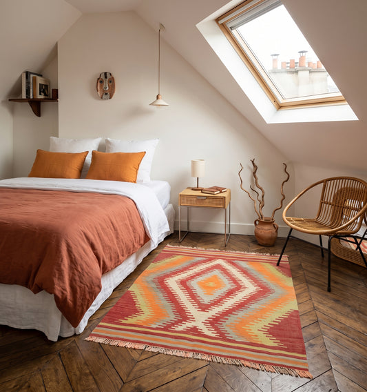 Petit tapis de salon en laine Cal aux motifs de losanges ocre et rouge (134 x 115 cm) mis en situation avec un lit dans une chambre à Paris avec déco à l'ambiance bohème en rotin.
