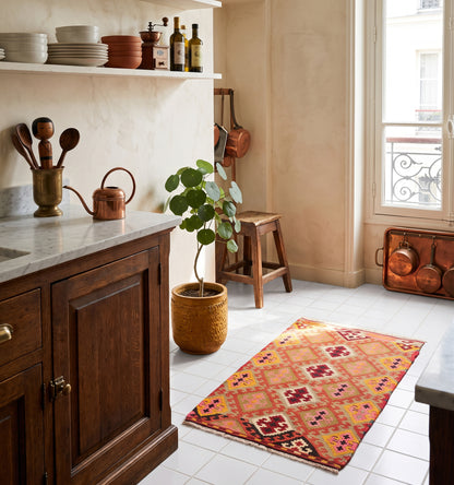 Tapis kilim ancien de Sivas, tissé main en laine par des nomades d'Anatolie, mosaïque de losanges rose et safran, posé sur carrelage blanc dans une cuisine parisienne au plan de travail en marbre et casseroles en cuivre