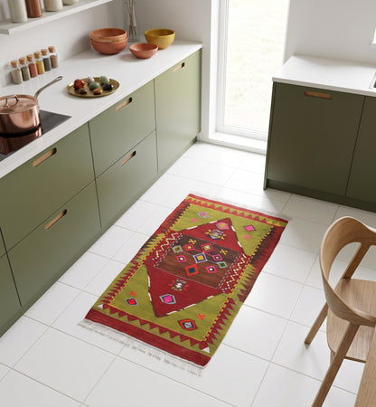 Tapis kilim ancien de Yozgat, tissé main en laine par des nomades d'Anatolie, médaillon losangé rouge et brun sur bordure dents de loup vert olive, posé dans une cuisine nantaise aux façades vert olive et carrelage blanc