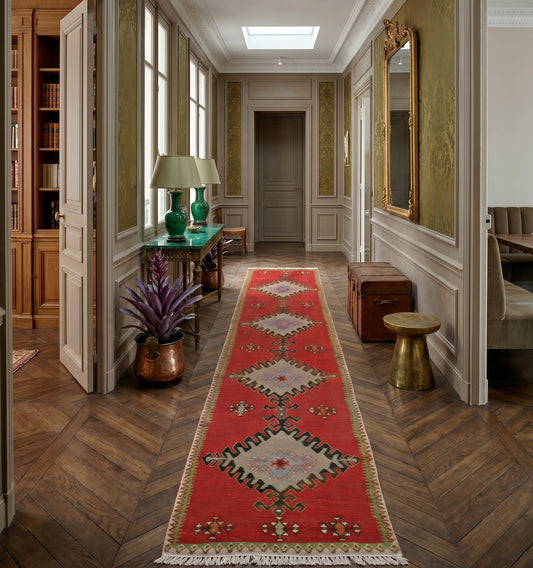 Tapis de couloir kilim ancien d'Anatolie rouge vif aux motifs de grands losanges, sublimant un long passage sur parquet chevronné à Versailles. Ce tissage artisanal traditionnel apporte un contraste chaleureux et chic au mobilier classique et aux moulures d'époque.