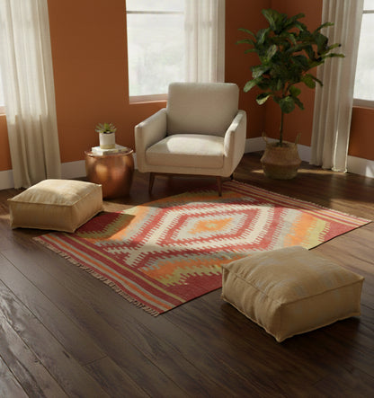 Petit tapis de salon en laine Cal aux motifs de losanges ocre et rouge (134 x 115 cm) mis en situation avec un fauteuil crème, des poufs de sol assortis et des murs de couleur terre cuite
