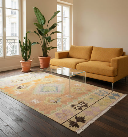 apis de salon en laine Cal aux teintes ocre, pastel et motifs géométriques (215 x 134 cm) mis en situation dans un intérieur moderne avec canapé jaune moutarde, table basse transparente en verre, grandes plantes vertes et parquet sombre
