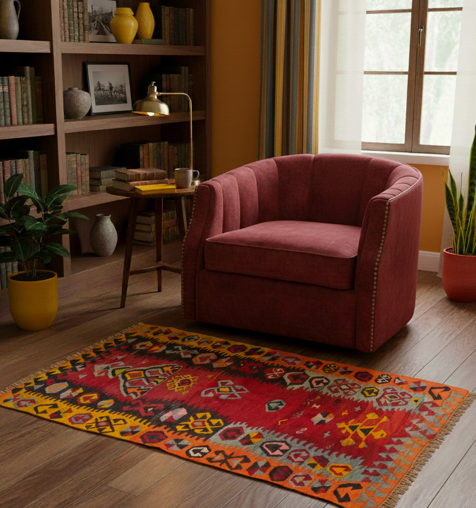 Petit tapis de salon en laine rouge et jaune aux motifs ethniques (145 x 90 cm) mis en situation devant un fauteuil en velours bordeaux et une bibliothèque